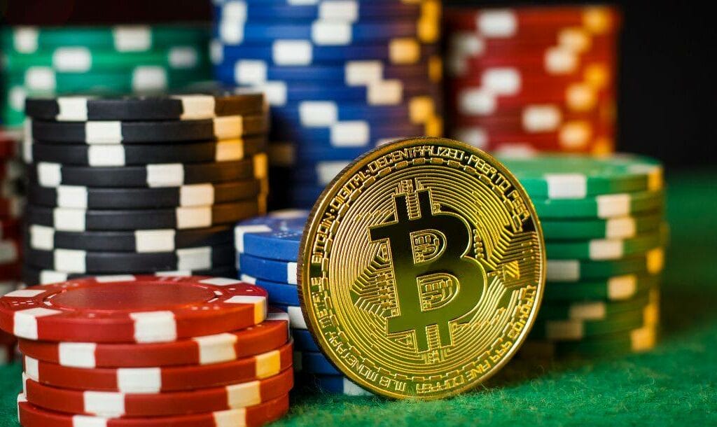 A gold Bitcoin balancing against multiple stacks of poker chips on a green felt table.