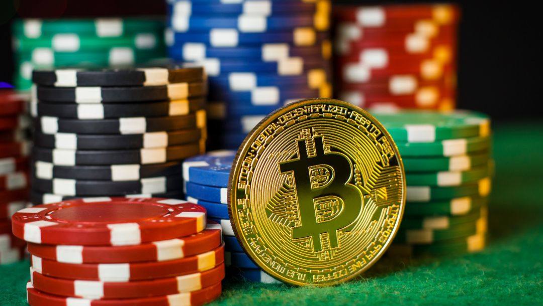 A gold Bitcoin balancing against multiple stacks of poker chips on a green felt table.