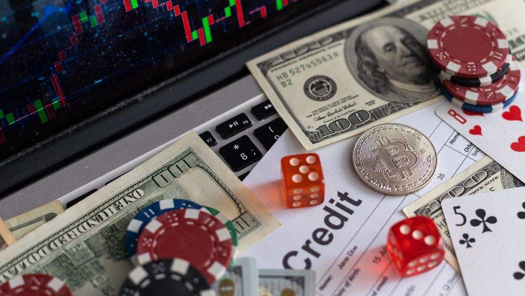 A laptop showing a chart on the screen and poker chips, cash, financial forms, dice, and a silver Bitcoin on the keyboard.