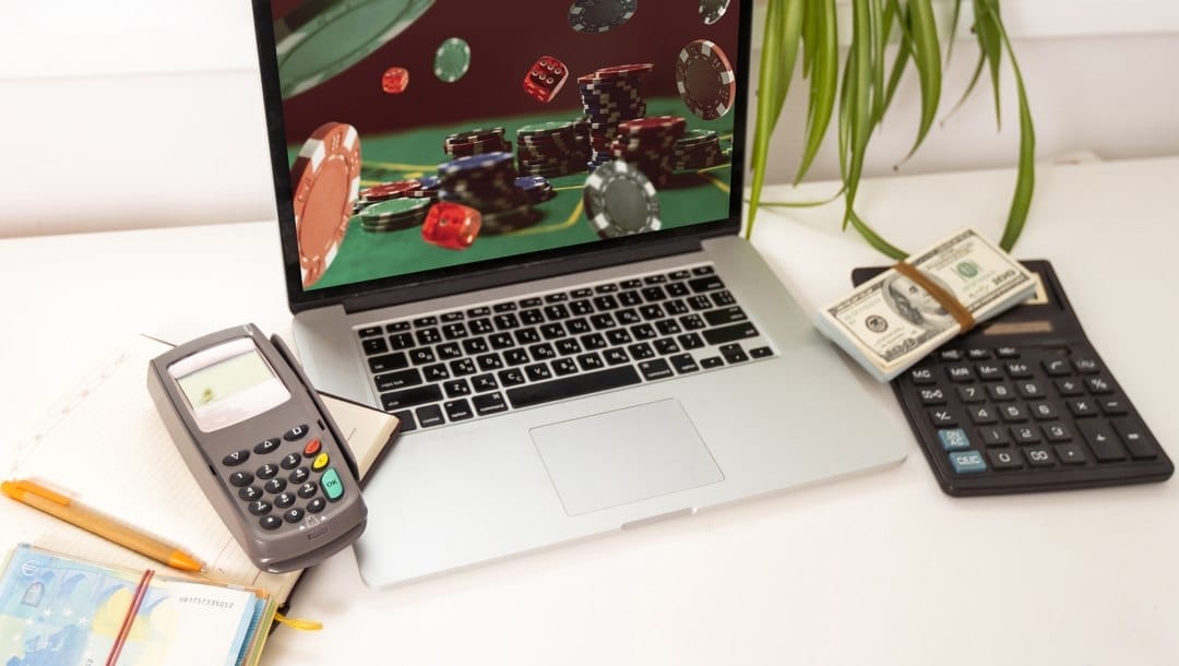 A laptop with poker chips on the screen is surrounded by calculators, a stacks of banknotes, and a notebook and pen
