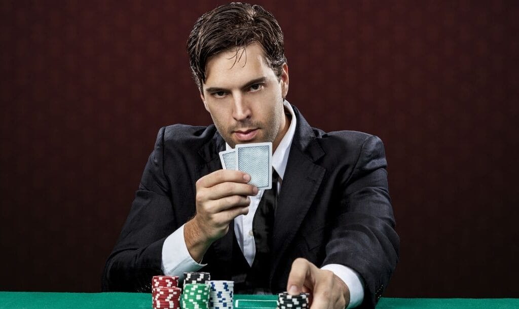 A man wearing a suit, with a very serious expression on his face, seated at a poker table, holding two playing cards in one hand, pushing poker chips forward with the other hand.