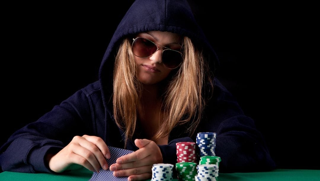 A woman wearing a hoodie and sunglasses, seated at a poker table, holding playing cards, with a few stacks of poker chips in front of her.