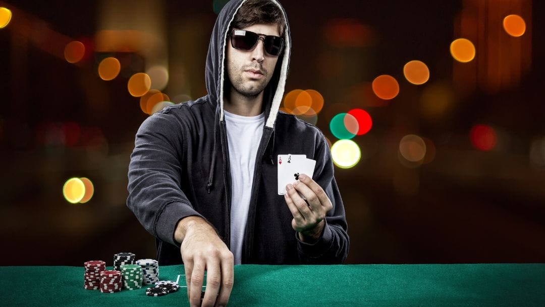 A man wearing sunglasses and a hoodie, seated at a poker table, holding two playing cards in one hand, and picking up poker chips with his other hand.