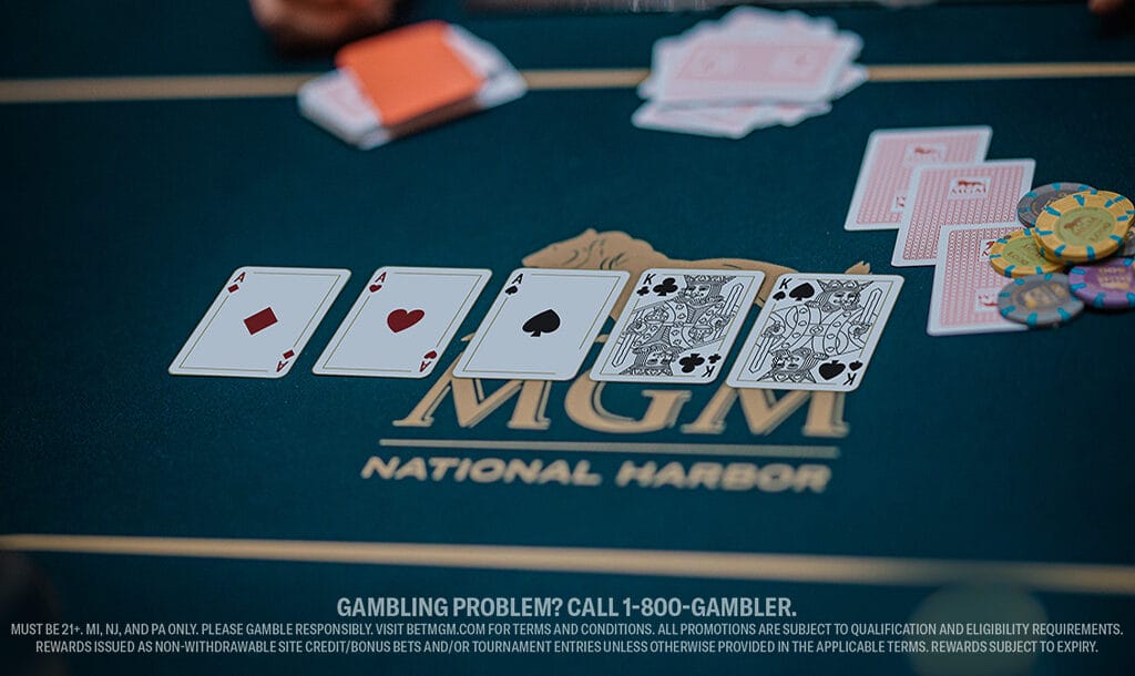 Poker table displaying five playing cards: ace, three, five of hearts, and face cards. Poker chips and cards lie on green surface with MGM logo.