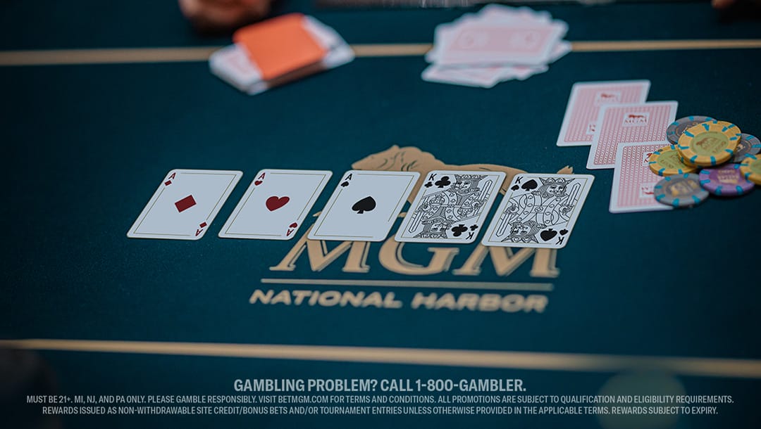 Poker table displaying five playing cards: ace, three, five of hearts, and face cards. Poker chips and cards lie on green surface with MGM logo.