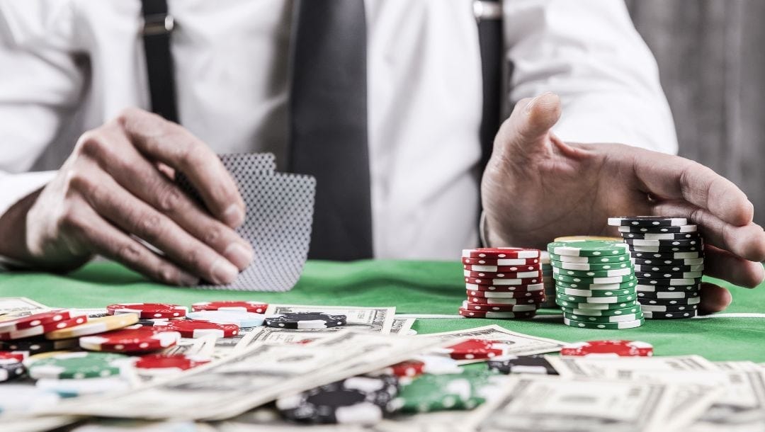 A person pushing poker chips forward with his one hand, and holding playing cards in their other hand, with poker chips, and cash arranged on top of the table.