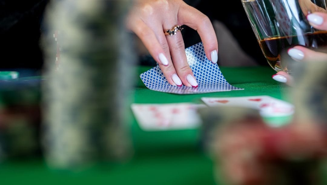 A person putting two playing cards down on a poker table, with drinks, poker chips, and playing cards arranged on top of the table.