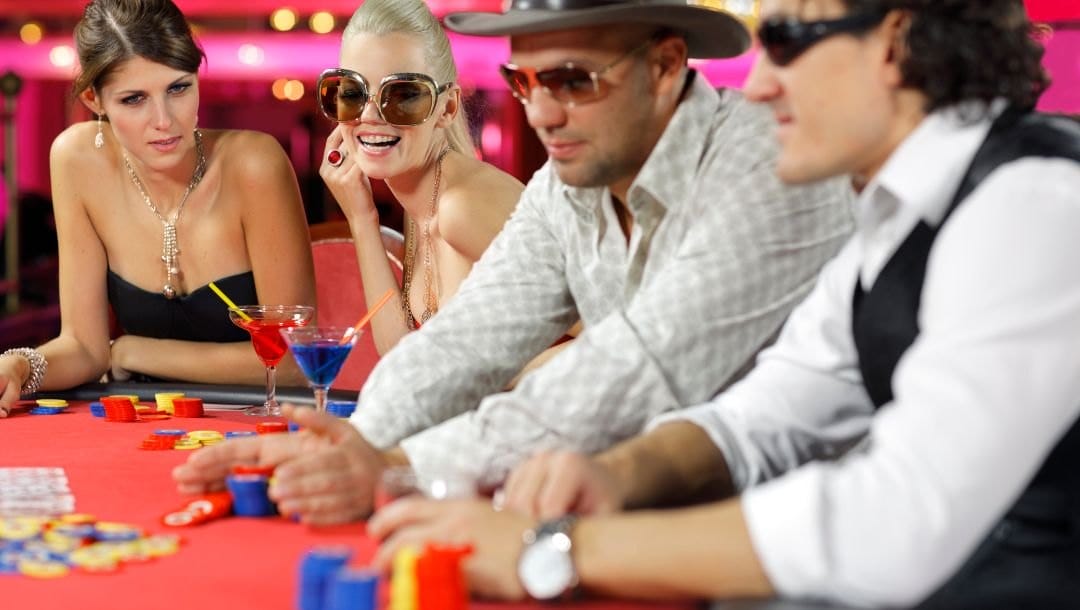 At a poker table, two women focus on the first man pushing chips, one intense, the other smiling, while the second man holds a drink.