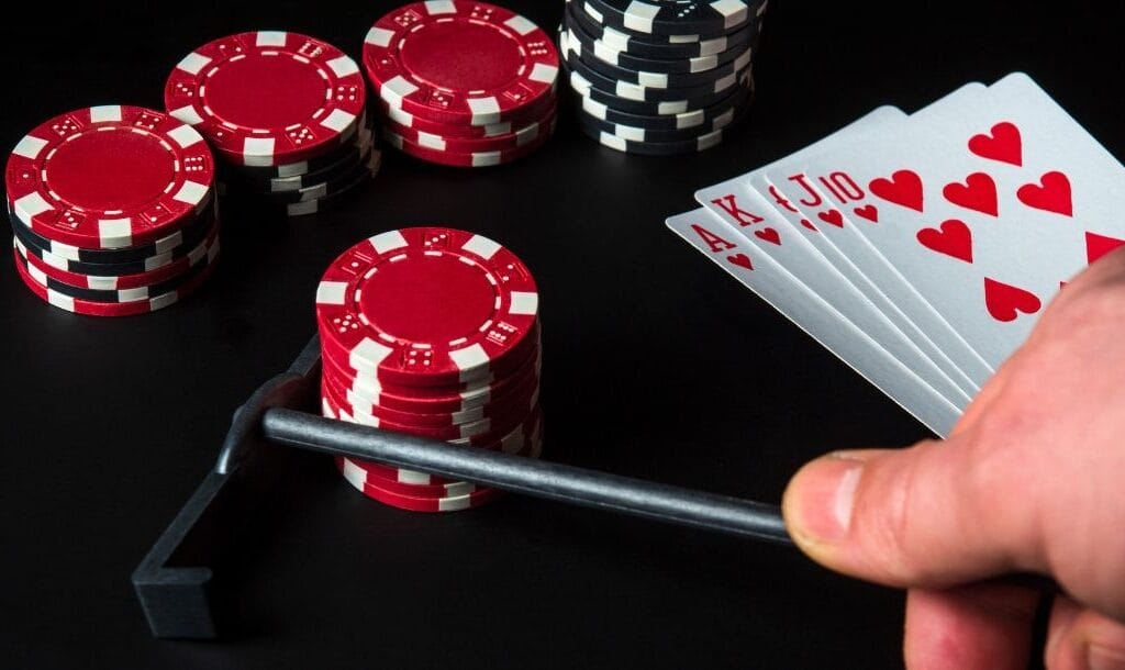 Person using a mini rake pulls in red poker chips. Four more stacks in background, and cards: Ace, King, Queen, Jack, 10 of Hearts on the right.