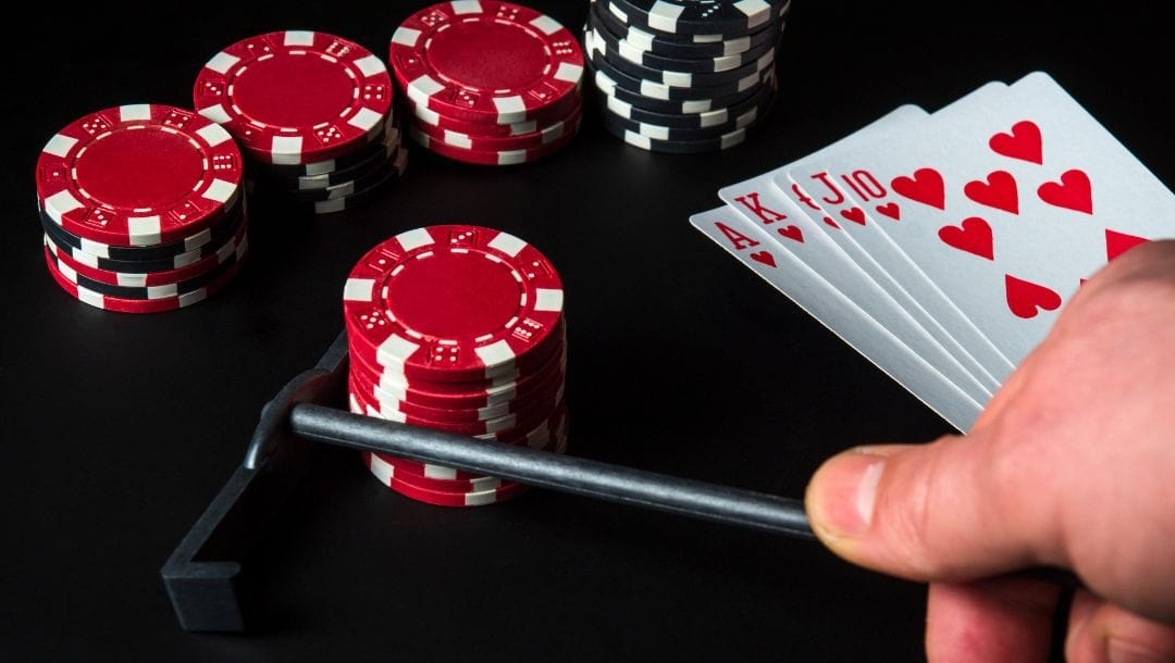 Person using a mini rake pulls in red poker chips. Four more stacks in background, and cards: Ace, King, Queen, Jack, 10 of Hearts on the right.