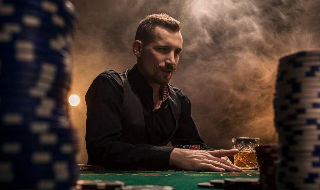 a man is sitting at a poker table with poker chips and a drink in front of him and smoke in the background, the shot is taken through two towers of poker chips at a low angle