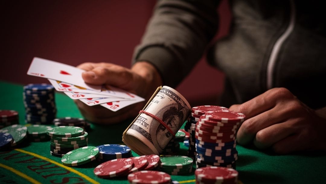 A person sitting at a poker table, holding cards, with poker chips and a roll of money arranged in front of them.