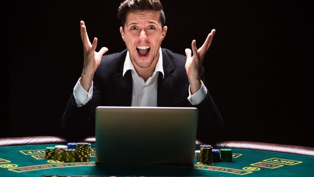 A very excited man in a suit sits at a poker table cheering with his laptop and stacks of poker chips in front of him.
