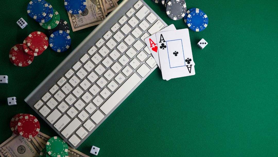 A wireless keyboard surrounded by playing cards, dice, poker chips, and cash on a green felt poker table.