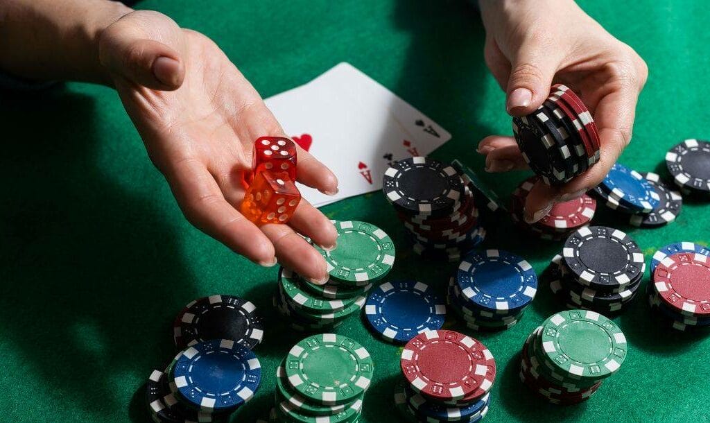 a person holds out a small stack of poker chips and a pair of red dice over a green felt poker table with poker chips and playing cards on it