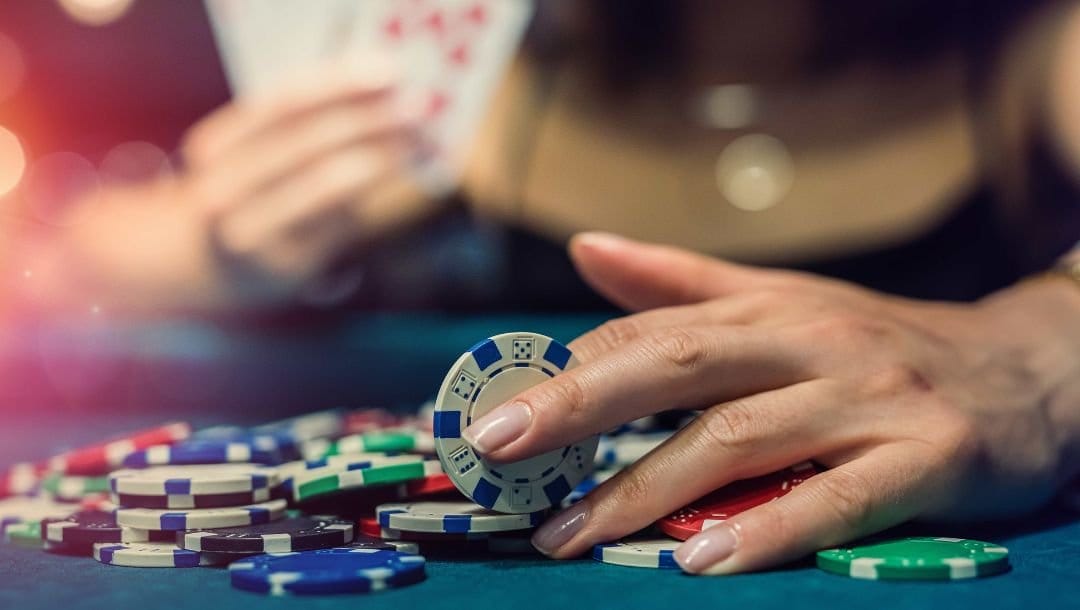 a woman holding a poker chip over a pile of poker chips on a blue felt casino table, she is holding playing cards up in the blurred background