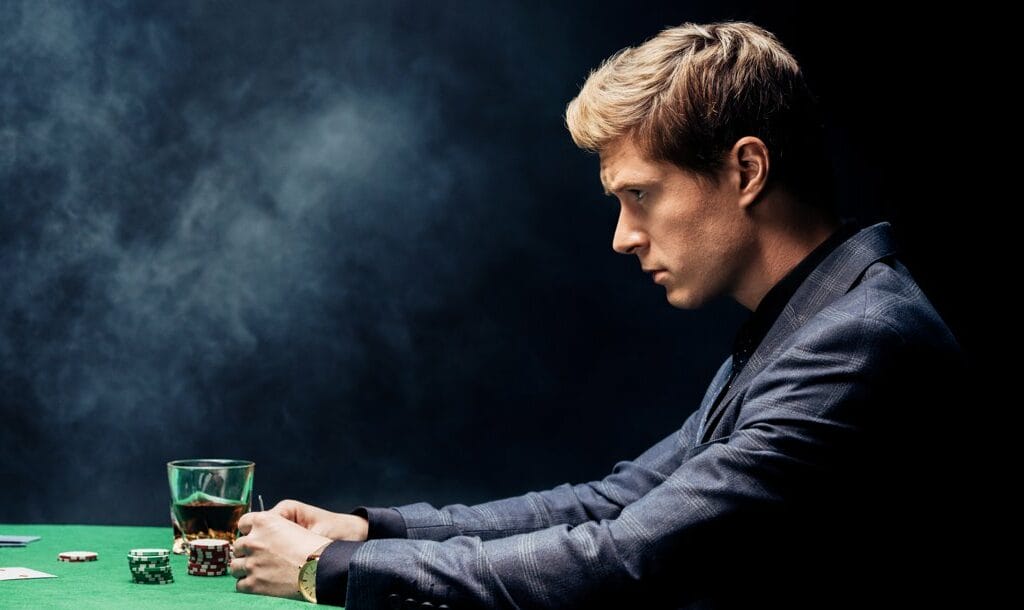 Side-view of a poker player in a suit sitting at a poker table. They are looking straight ahead with an expressionless face.
