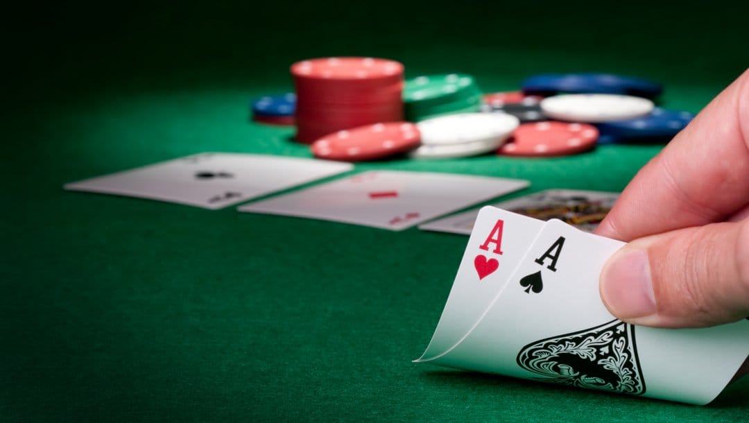 A poker player checks their hole cards. They have two aces. The community cards and pot are in the background.