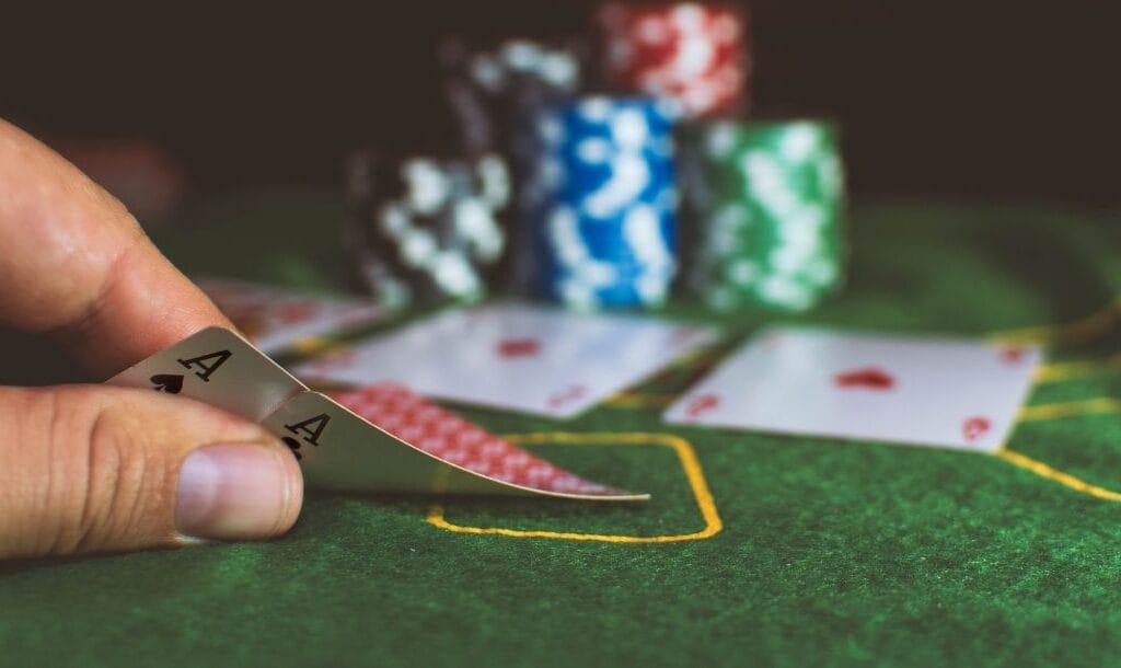 Hand lifting two cards to reveal an ace of clubs and an ace of spades. Stacks of poker chips in the blurred background.