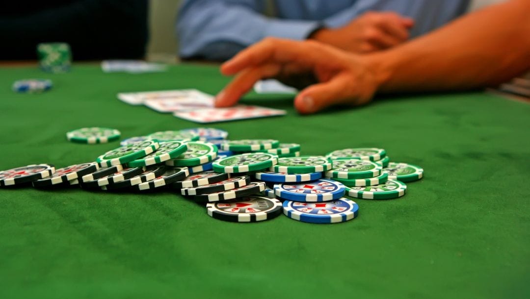 Poker chips scattered around on a green felt table. Hand in the background fanning out three cards.