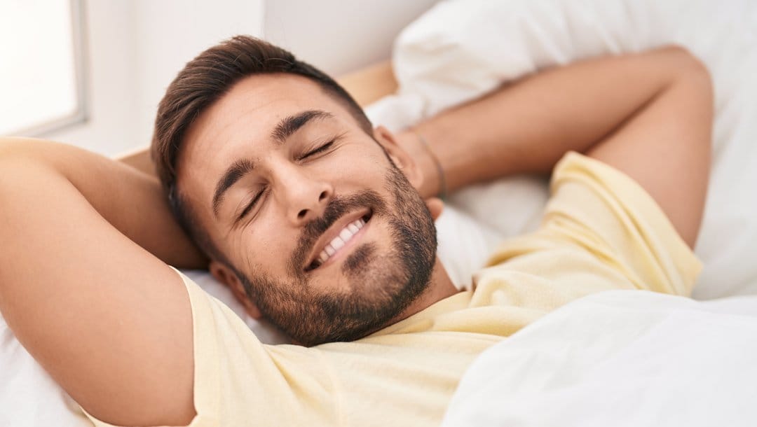 smiling person lying on a bed with their hands behind their head.