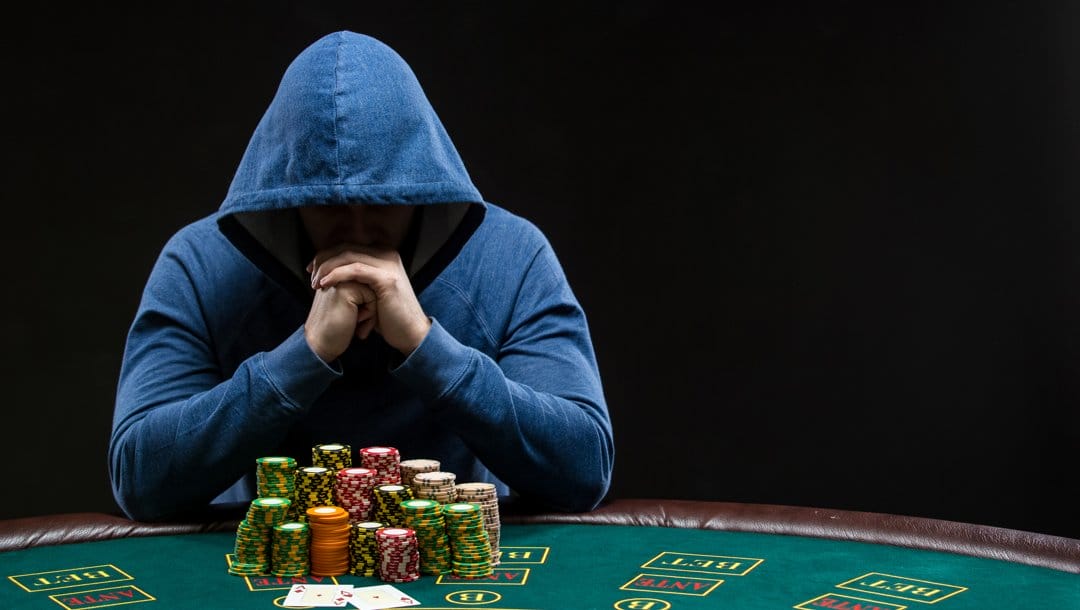 A poker player in a hoodie looking down with their hands covering their mouth. There are multiple large stacks of poker chips and two playing cards in front of them.