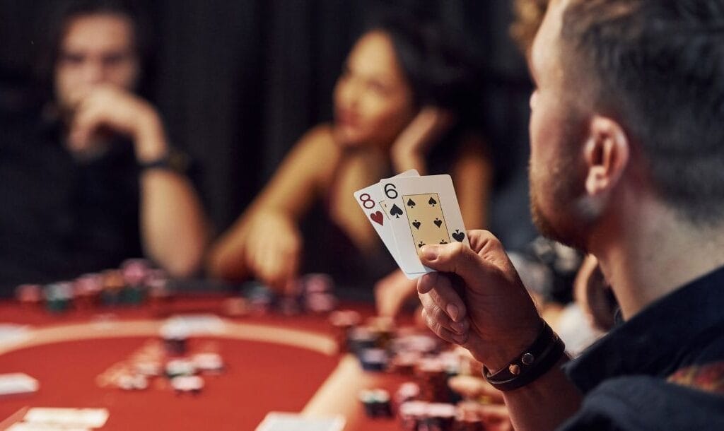 Table of poker players with focal point on a man holding an 8 of hearts and 6 of spades, other players are subtly blurred in the background.