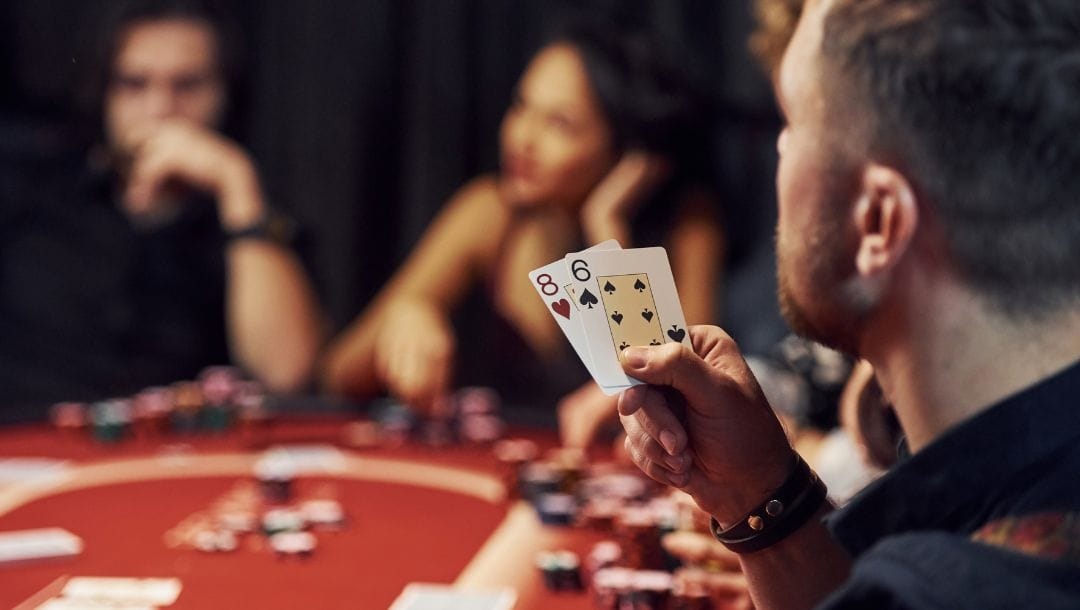 Table of poker players with focal point on a man holding an 8 of hearts and 6 of spades, other players are subtly blurred in the background.