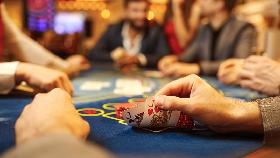 Person lifting up two cards to show a jack of hearts and a jack of clubs. Other players around the blue felt poker table are blurred in the background.