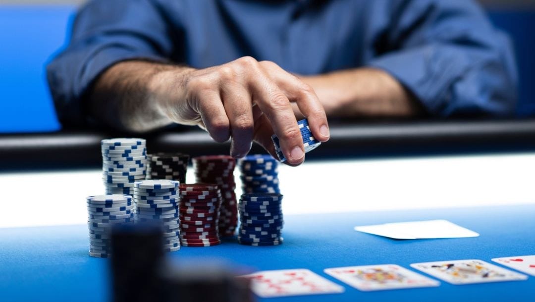 A man wearing a blue shirt is placing poker chips from eight stacks to the center of the blue felt table, with face-up cards laid out in front of him.