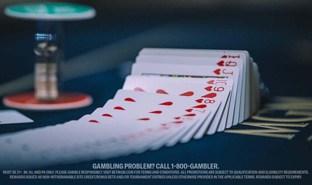 A deck of cards fanned out on a poker table, highlighting hearts. A poker chip and dealer button are blurred in the background.