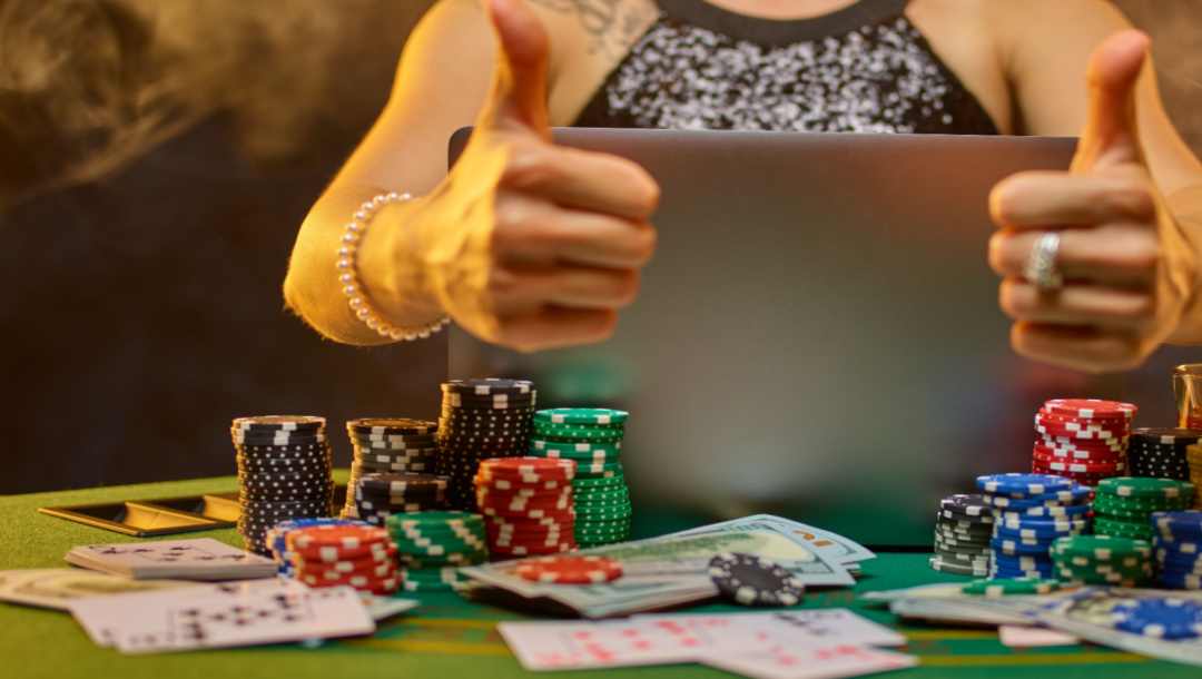 A person giving a thumbs-up gesture while using a laptop on a poker table. There are casino chips and cash on the poker table, too.