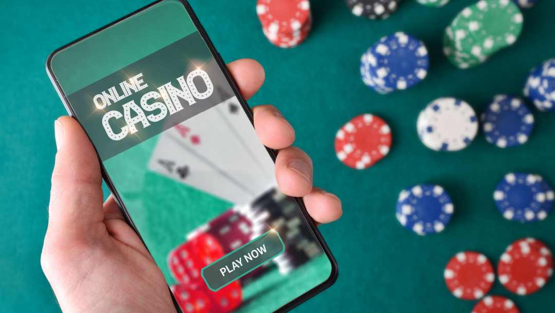 A person holding a smartphone with an online casino app open above a poker table where casino chips are spread out.