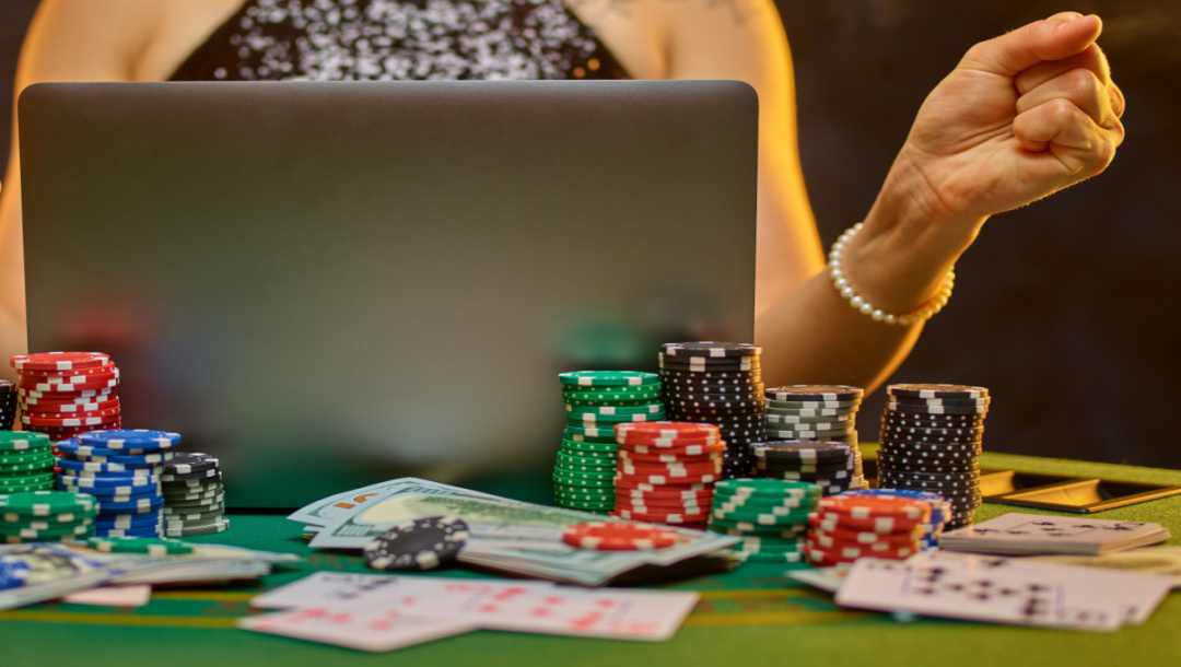 A person sits at a poker table with cash, chips, and cards surrounding them while they play on a laptop.
