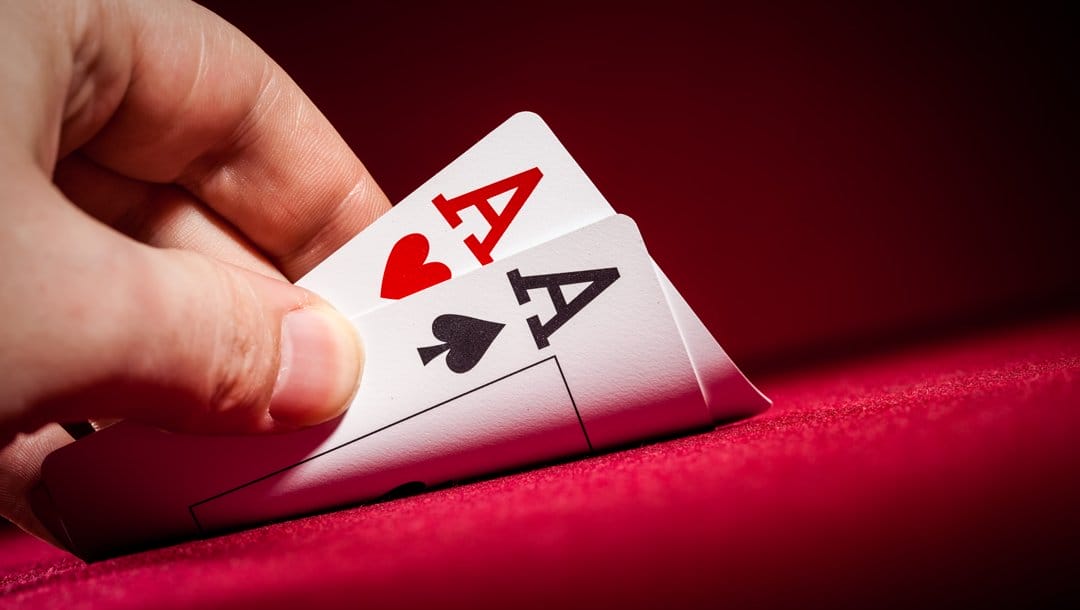 A player checks their hole cards, which lie face-down on a red casino table. They see a pair of aces.