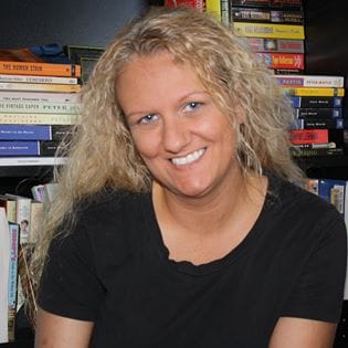 A smiling woman with curly hair, wearing a black top, in front of a bookshelf.