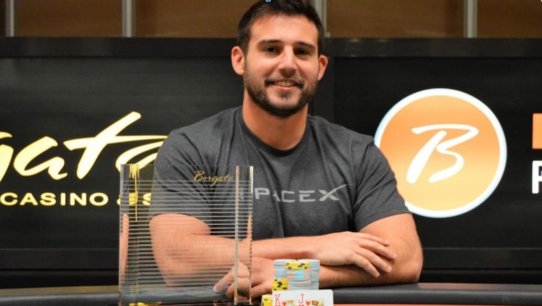 A man smiles at a table with a trophy and poker chips, at a casino.
