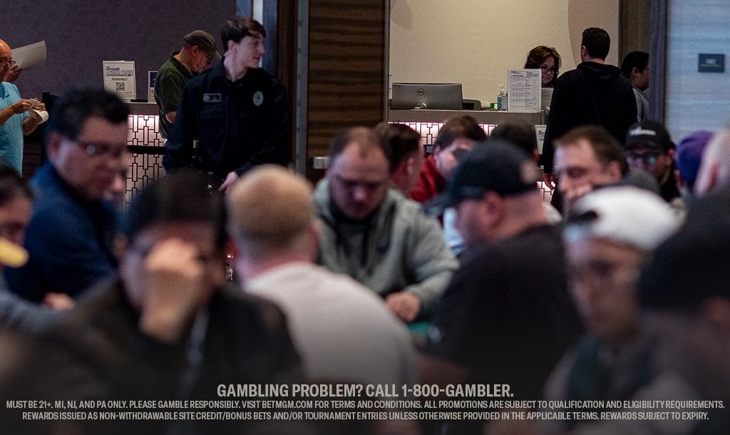 A busy poker room filled with focused players sitting around tables. A security guard stands in the background.