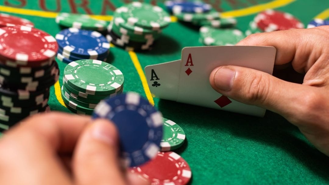 A person holding an Ace of Diamonds, and an Ace of Clubs in one hand, and a blue poker chip in the other hand, with poker chips arranged on a poker table.