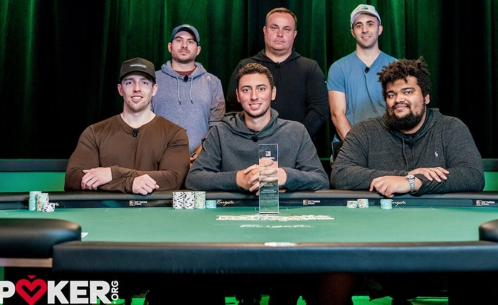 Six people pose around a poker table with chips; one holds a trophy, implying a tournament outcome.