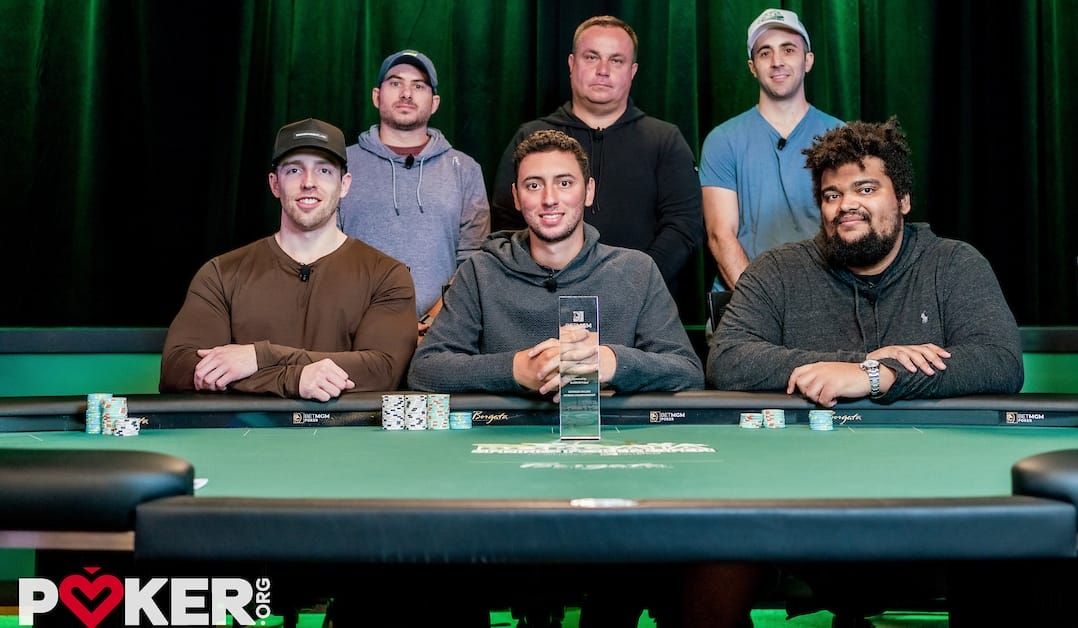 Six people pose around a poker table with chips; one holds a trophy, implying a tournament outcome.