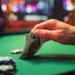 A hand holding playing cards at a poker table on a green felt table with casino chips.