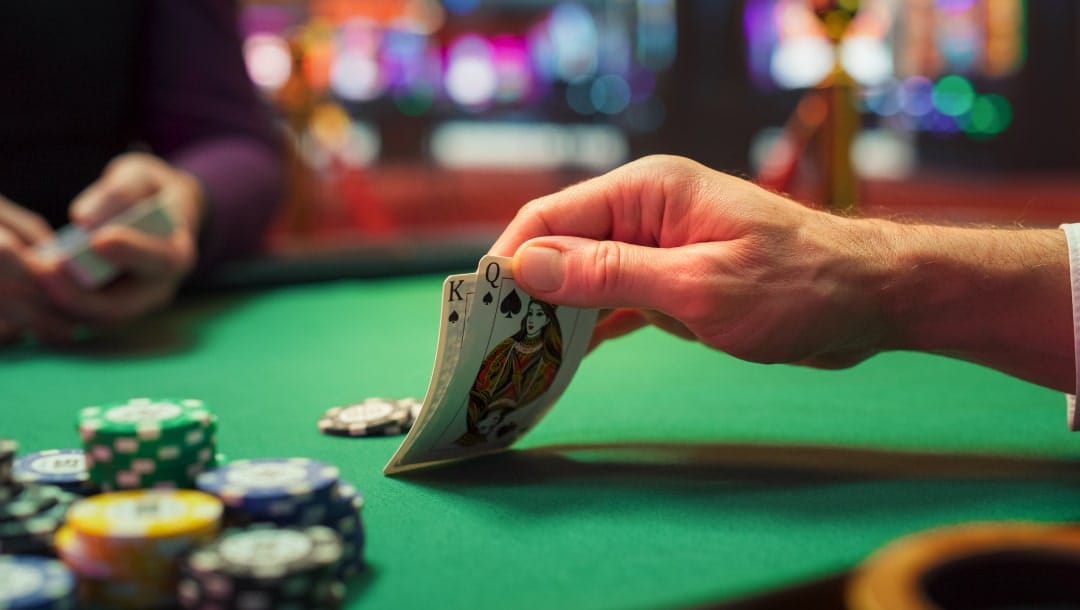 A hand holding playing cards at a poker table on a green felt table with casino chips.