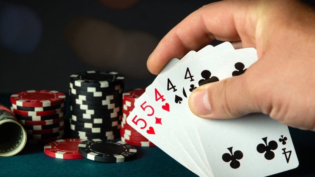 A hand holding playing cards with casino chips and a roll of money on a dark surface.