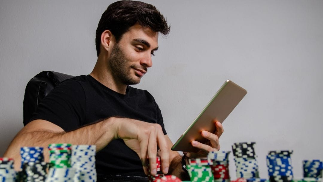 A man sitting at a table playing an online poker game on a tablet, with stacks of poker chips arranged on the table.