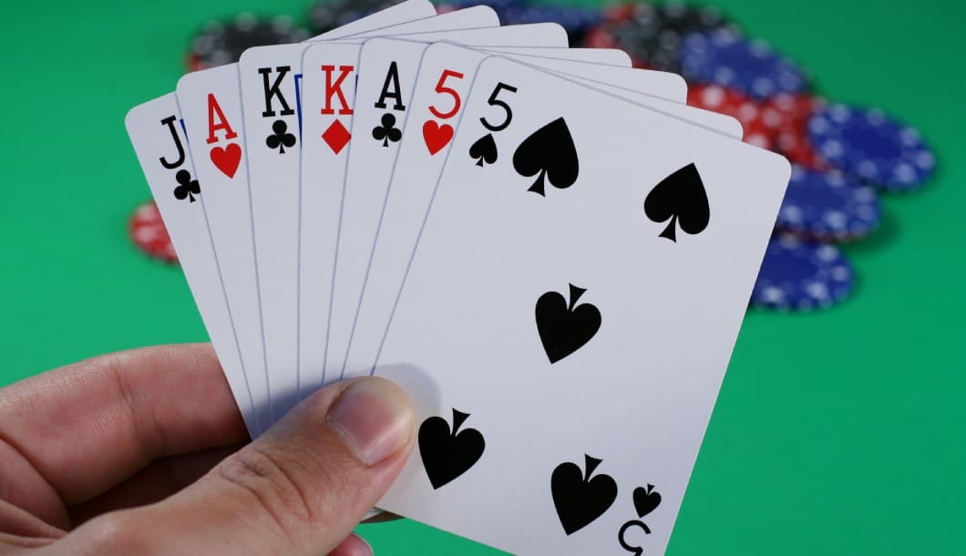 A hand holding seven playing cards in front of a green felt table with blue, red and black casino chips on the table.