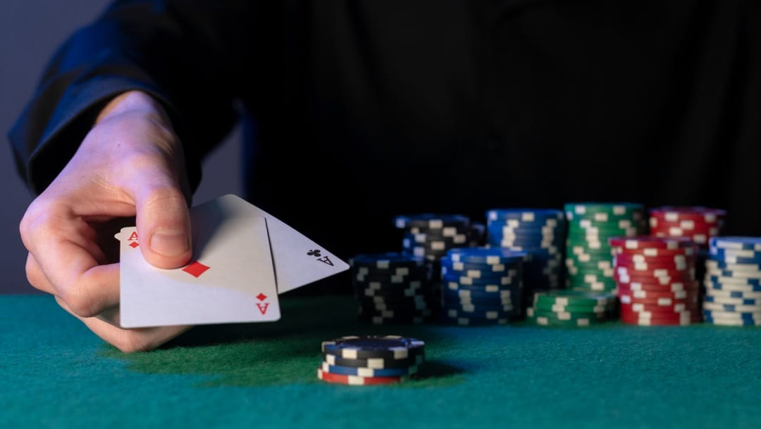 A hand holding two poker cards with red, blue, green, and white casino chips on a turquoise poker table.