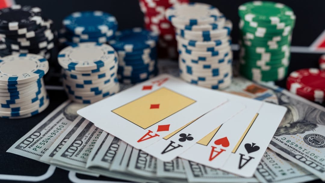 Four ace playing cards, 100 dollar bills, and red, black, white, and green casino chips on a black poker table.