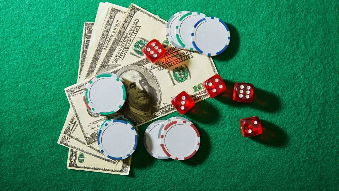 $100 Bills fanned out on a green felt table with poker chips and five red six-sided dice on top of them.