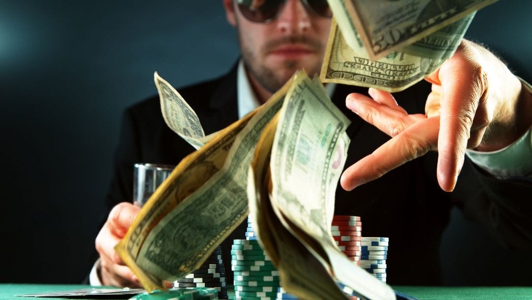 A poker player wearing sunglasses sitting at a poker table with stacks of chips in front of him throws money towards the camera.